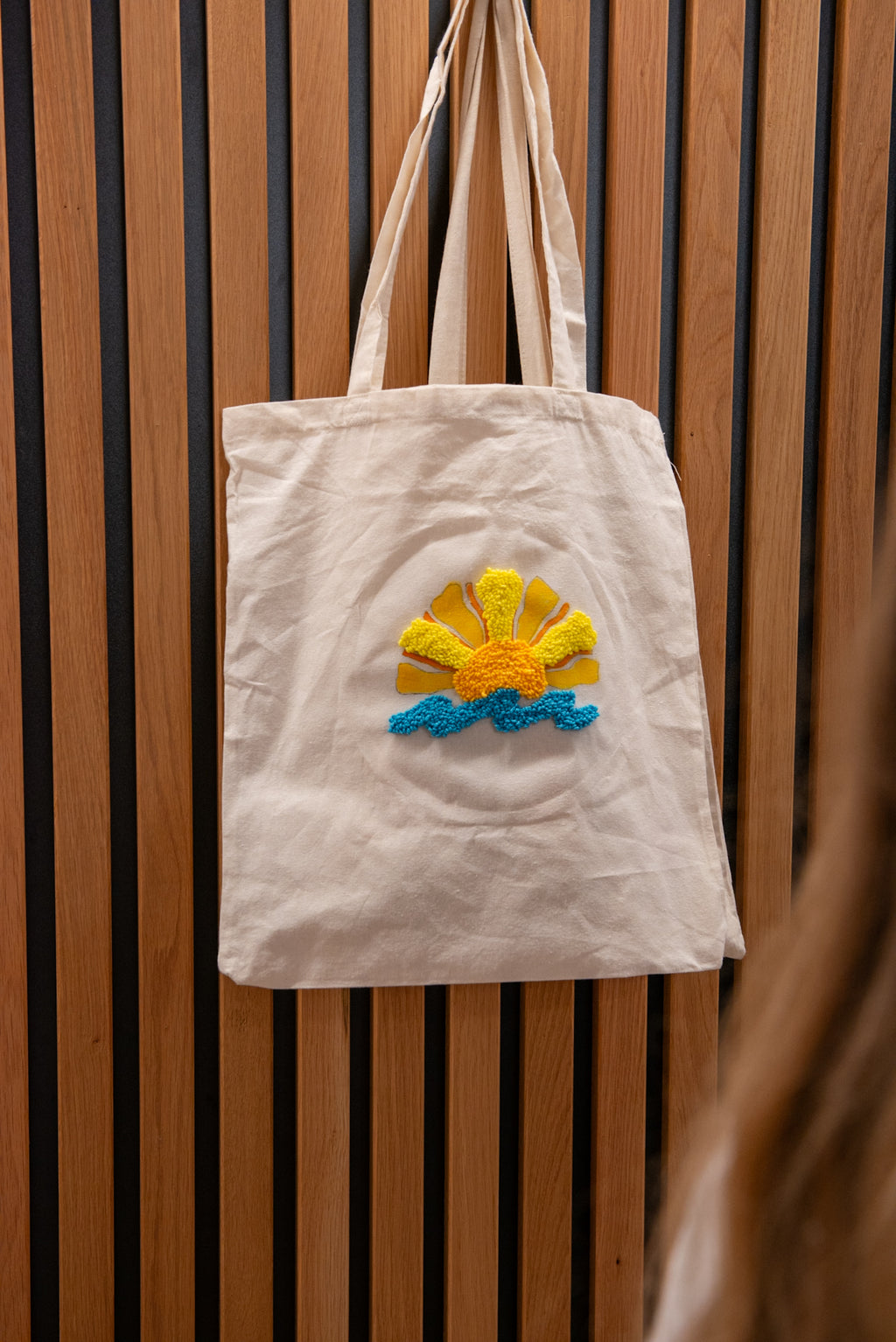 Eine „Straightcurls Jutebeutel FFB Emmering“-Tragetasche mit Sonnenmotiv hängt nach einem kreativen Kindergeburtstag an einer Holzwand.