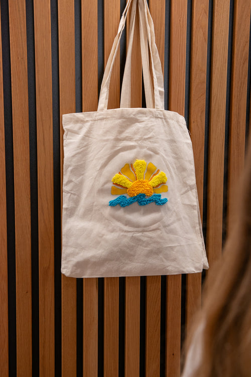 Eine „Straightcurls Jutebeutel FFB Emmering“-Tragetasche mit Sonnenmotiv hängt nach einem kreativen Kindergeburtstag an einer Holzwand.