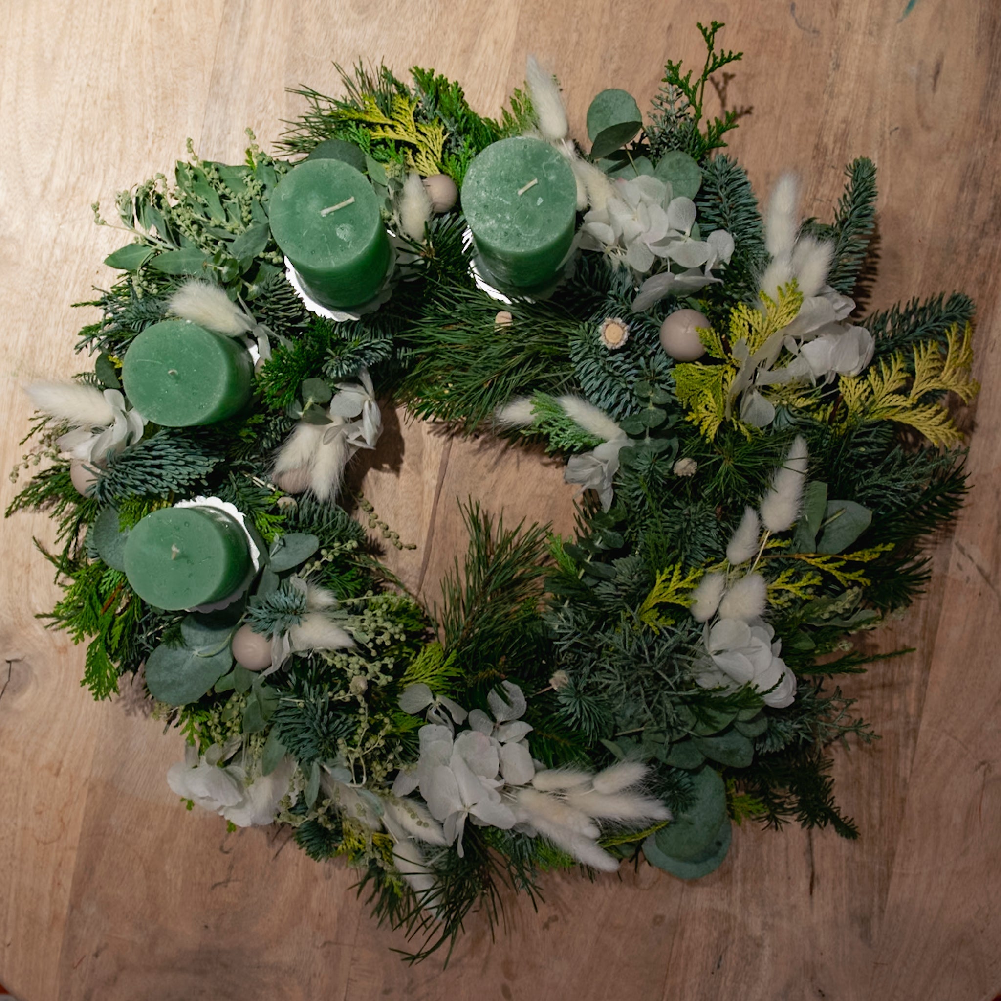 Ein StraightCurls-Adventskranz aus der FFB-Werkstatt in Emmering mit grünen Kerzen und weißen Blüten ruht auf einem Holzsockel.