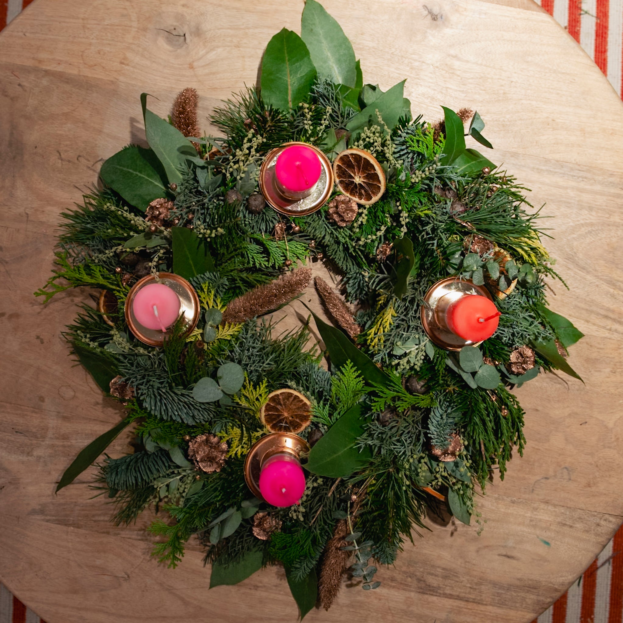 StraightCurls’ Adventskranz-Workshop FFB Emmering: grüner Kranz, 4 rosa Kerzen, Orangenscheiben, Tannenzapfen auf Holz.