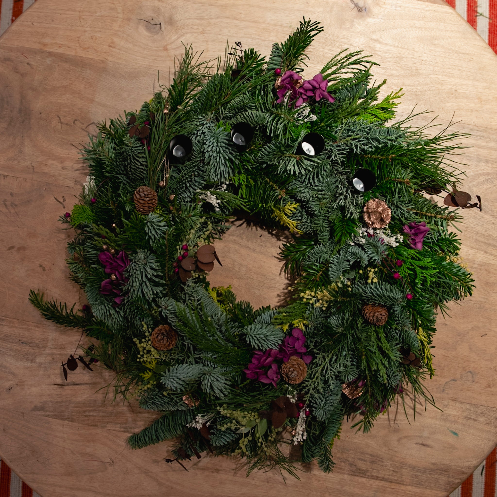 Der Adventskranz aus dem StraightCurls-Workshop FFB Emmering mit Tannenzapfen, Blumen und Kerzen steht auf einem Tisch.