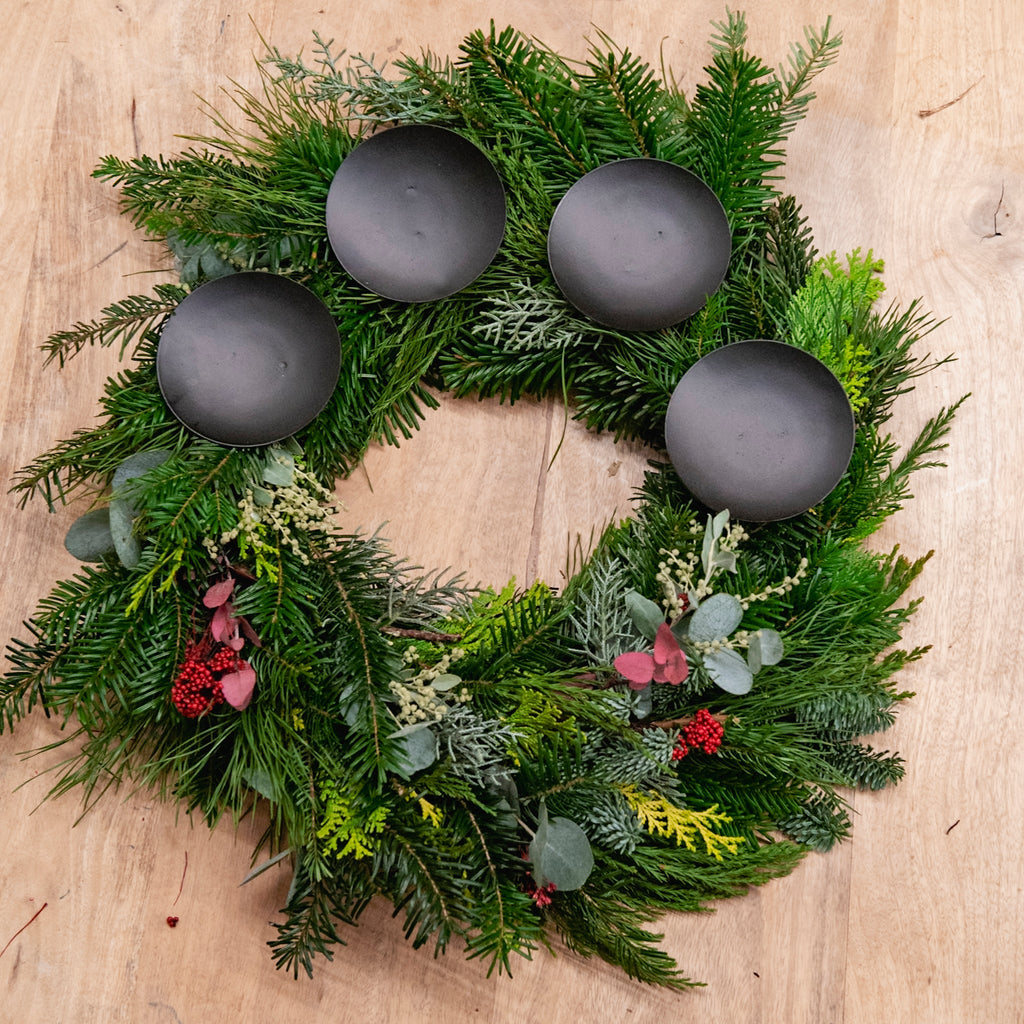 Ein StraightCurls-Adventskranz-Workshop: Ein FFB-Emmering-Kranz mit 4 schwarzen Haltern und roten Beeren auf einem Holzsockel.