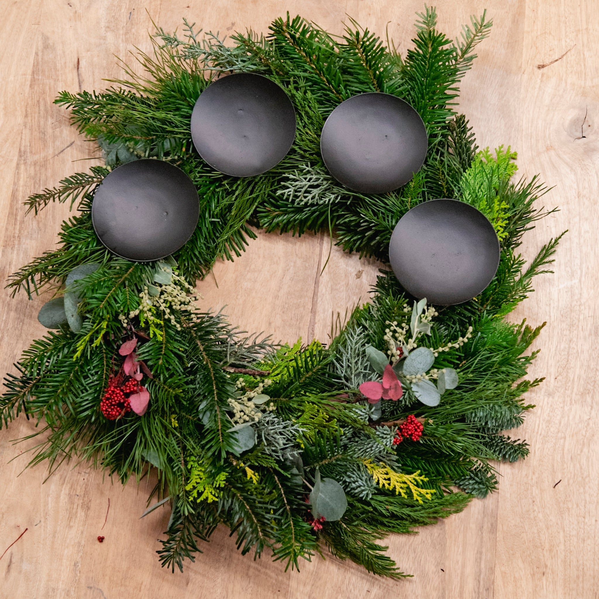 Ein StraightCurls-Adventskranz-Workshop: Ein FFB-Emmering-Kranz mit 4 schwarzen Haltern und roten Beeren auf einem Holzsockel.