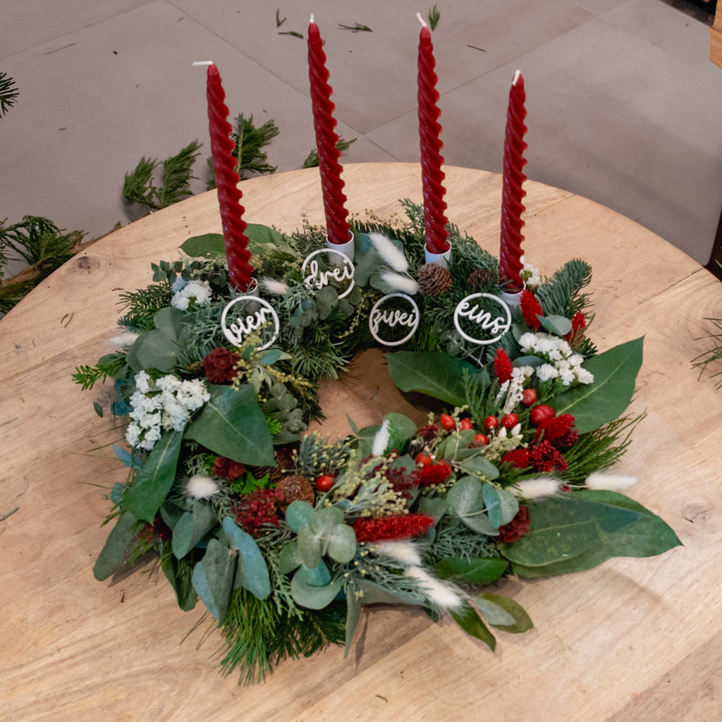 StraightCurls-Adventskranz-Workshop FFB Emmering: Vier rote Kerzen, Tannengrün, auf einem Holztisch mit den Nummern 1–4 beschriftet.