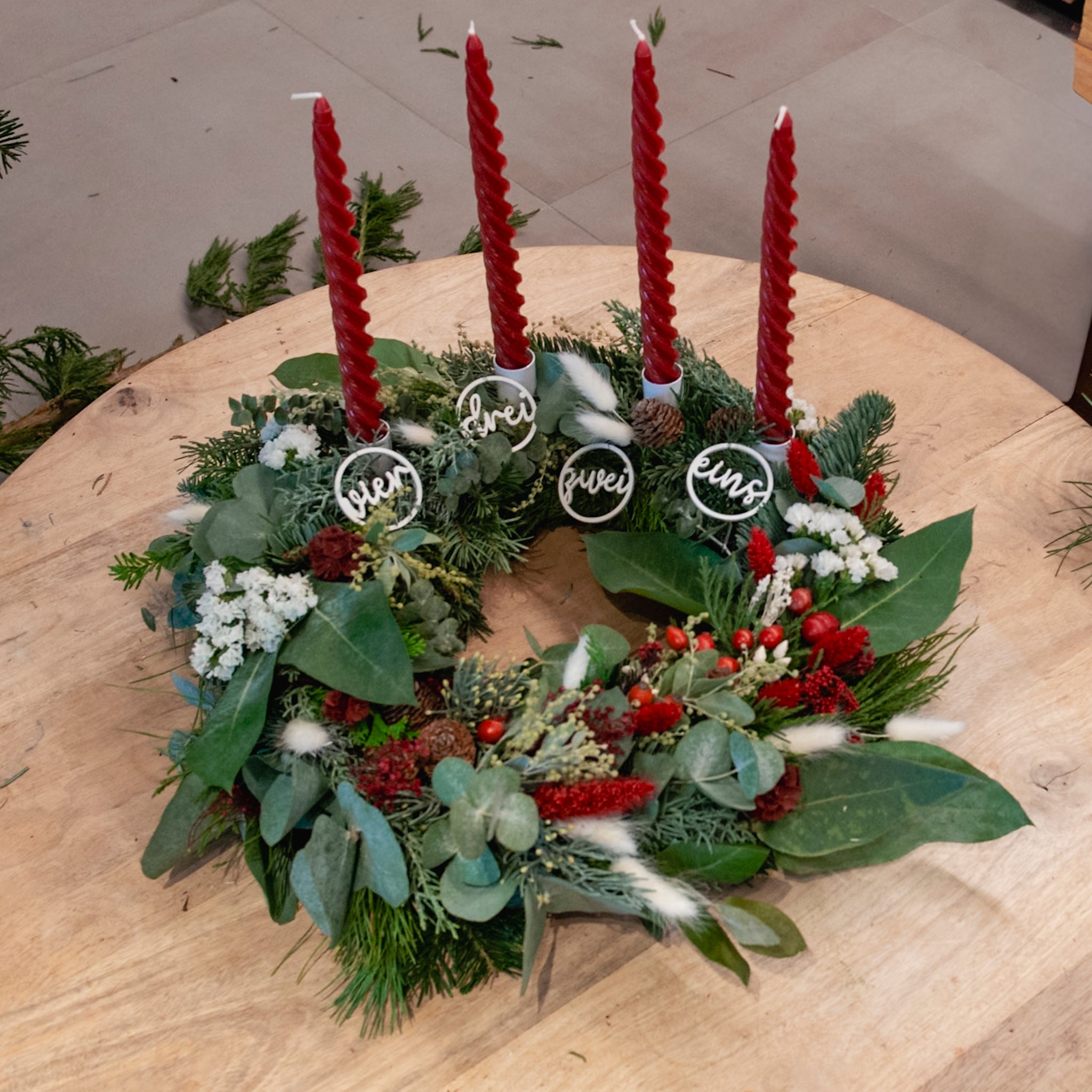 StraightCurls-Adventskranz-Workshop FFB Emmering: Vier rote Kerzen, Tannengrün, auf einem Holztisch mit den Nummern 1–4 beschriftet.