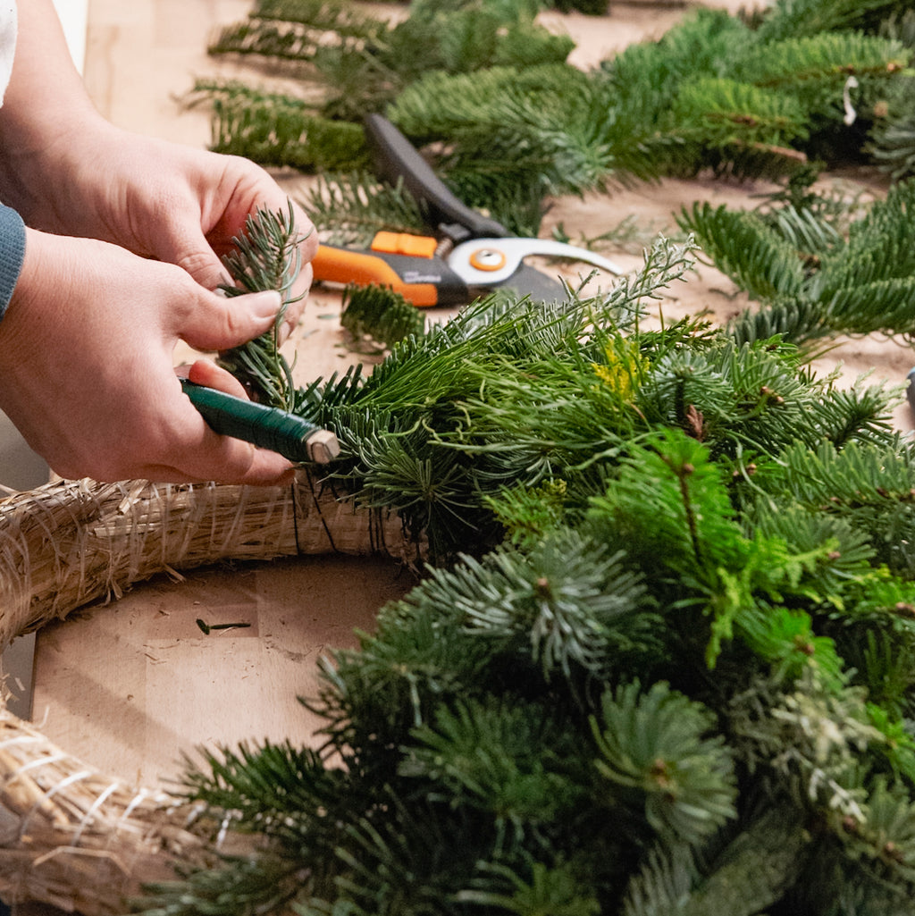 Beim StraightCurls-Adventskranz-Workshop in FFB Emmering befestigen Teilnehmer Tannengrün an einem Kranz; daneben liegen Scheren.