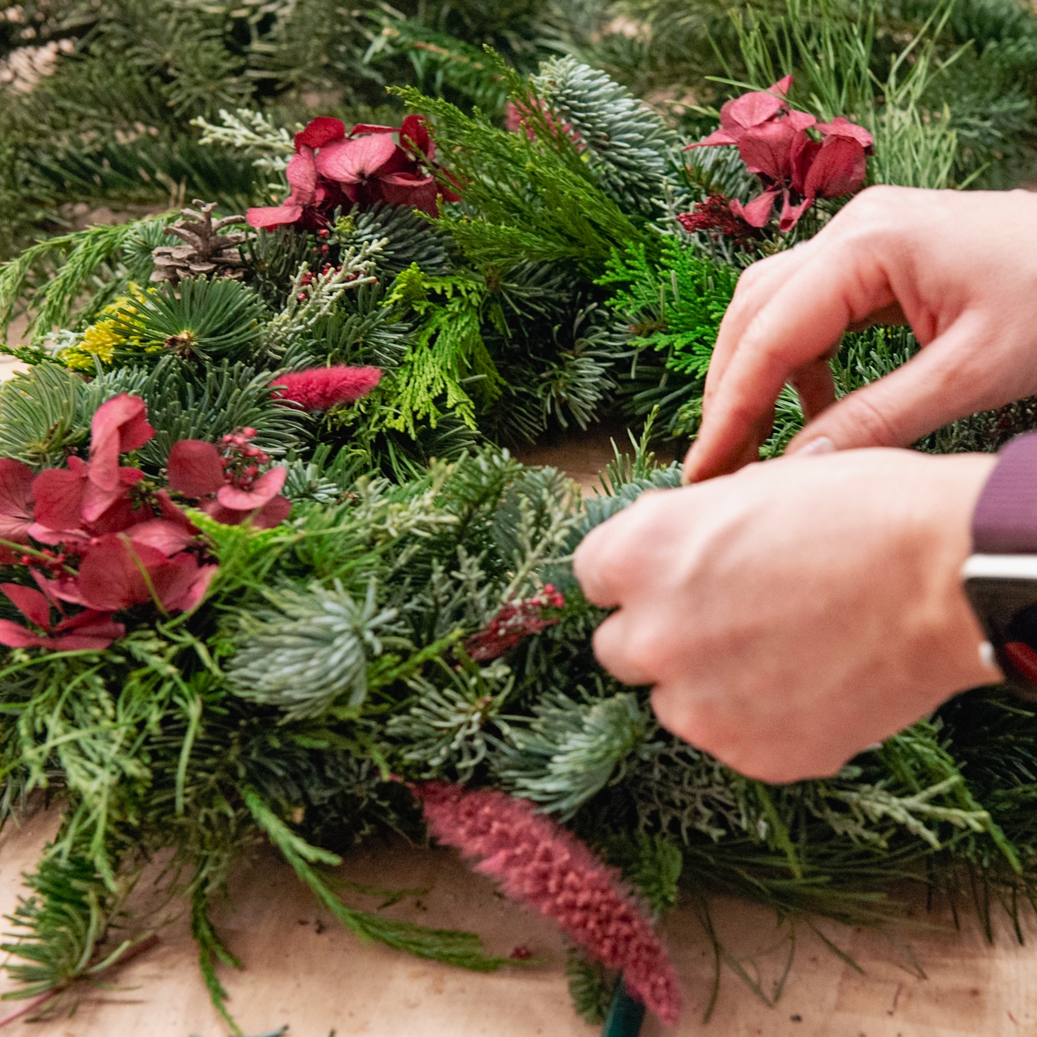 Hände, die bei einem Adventskranz-Workshop von StraightCurls in FFB Emmering einen Adventskranz mit roten Blüten basteln.