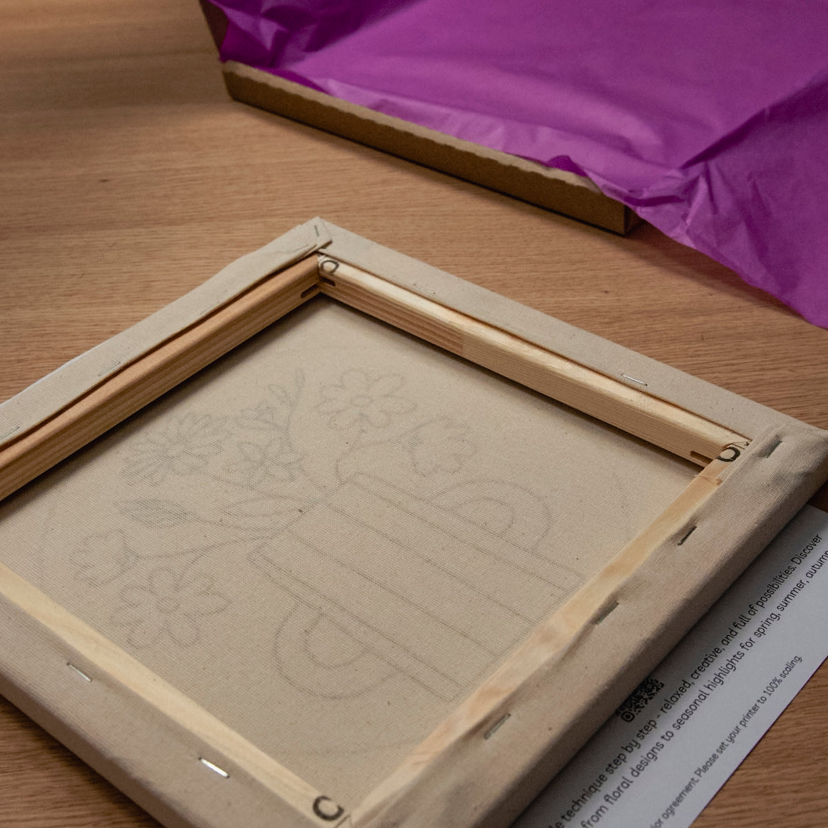 Neben einer Leinwand mit Blumenmotiv liegen ein „Straightcurls Punch Needle“-Keilrahmen (24 × 24 cm) und lila Seidenpapier.