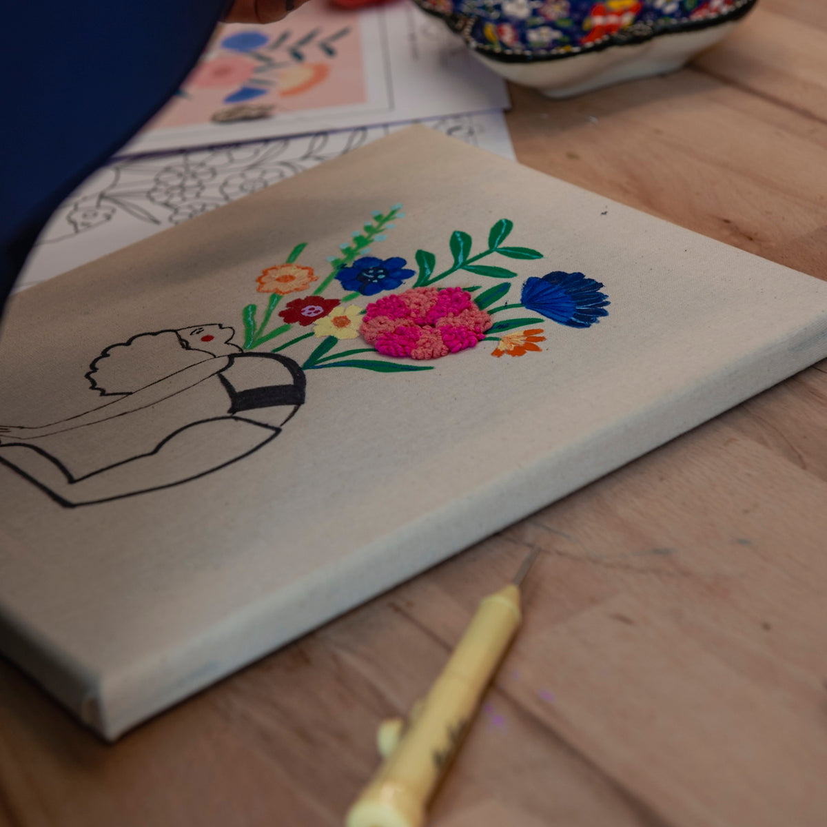 „Straightcurls Punch Needle Keilrahmen 24 x 30 cm“ zeigt eine unvollständige Figur mit bunten Blumen und Malutensilien.