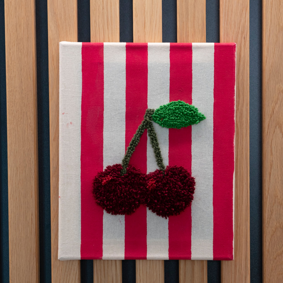 Straightcurls Punch Needle Keilrahmen 24 x 30 cm: rot-weiß gestreifter Baumwollstoff, Kirschmotiv, Holzplatte als Hintergrund.