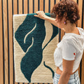 Eine Person hängt einen StraightCurls Tufting Workshop FFB Emmering-Teppich mit Grün- und Cremetönen an eine Lattenwand.