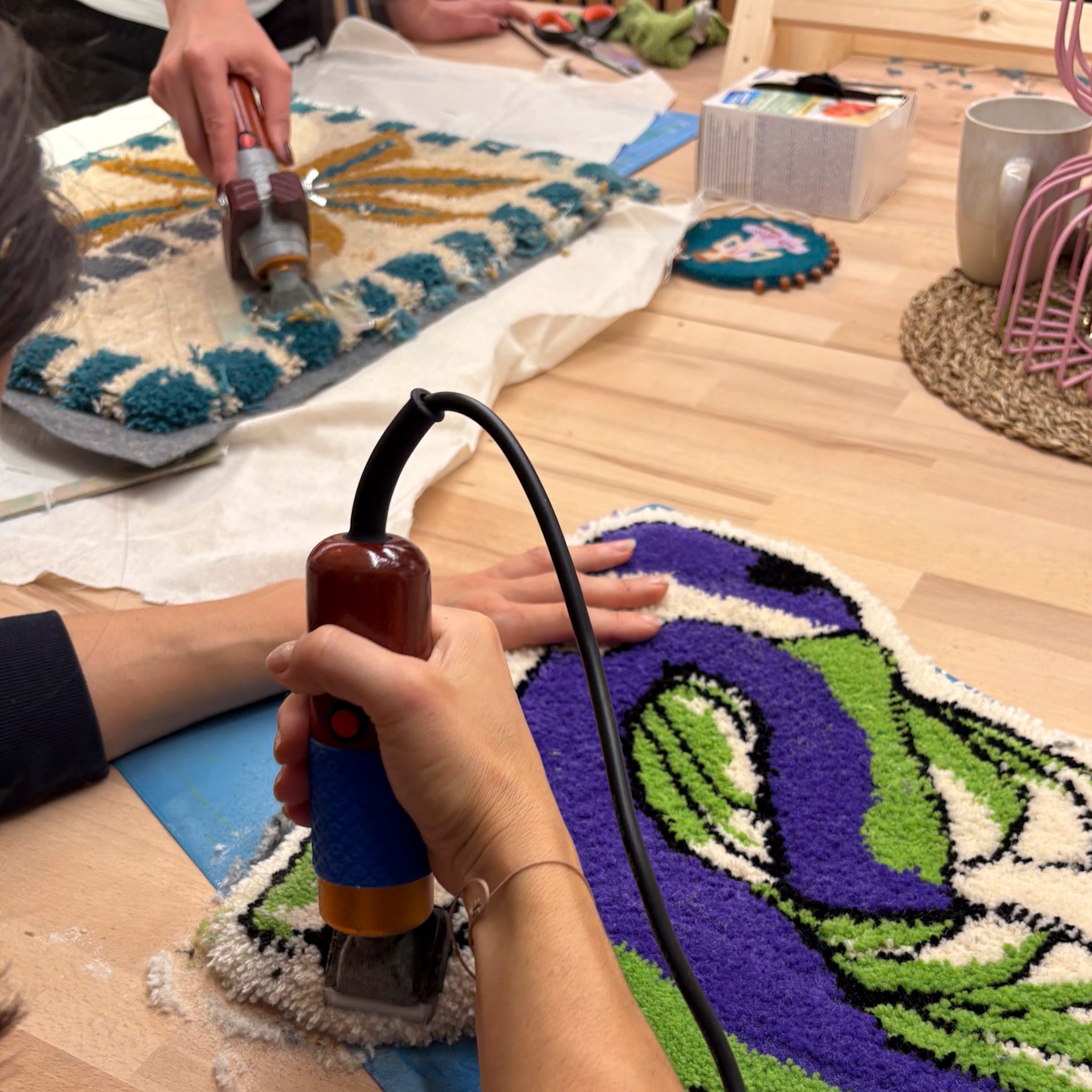 In der StraightCurls Tufting-Werkstatt FFB Emmering tuften zwei Personen auf einem Holztisch Teppiche aus bunten Materialien.