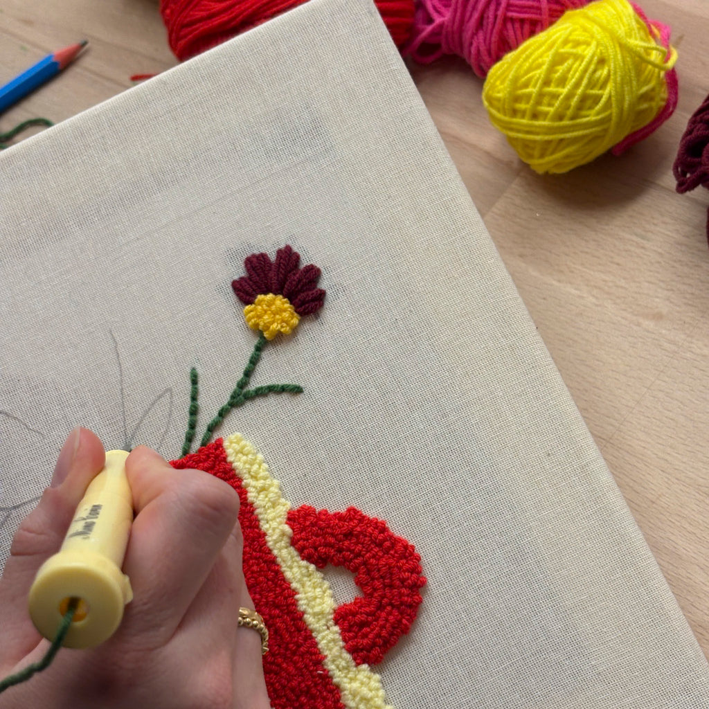 Eine Hand benutzt eine Stanznadel in einem StraightCurls Punch Needle Workshop FFB Emmering, um florale Stoffkunst mit Garn herzustellen.
