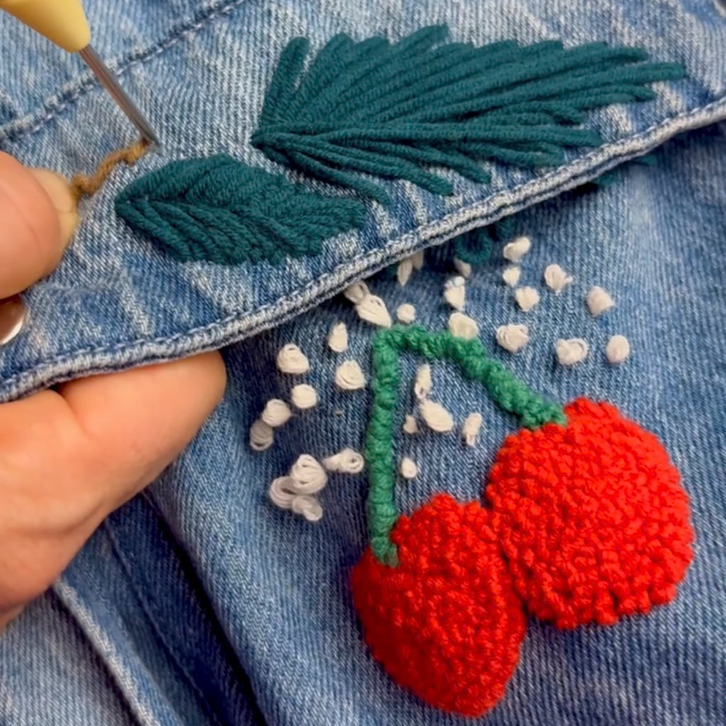 Detailaufnahme: Kirsch-Motiv wird mit der Stanznadel direkt auf eine Jeansjacke aufgebracht (Textil-Upcycling).