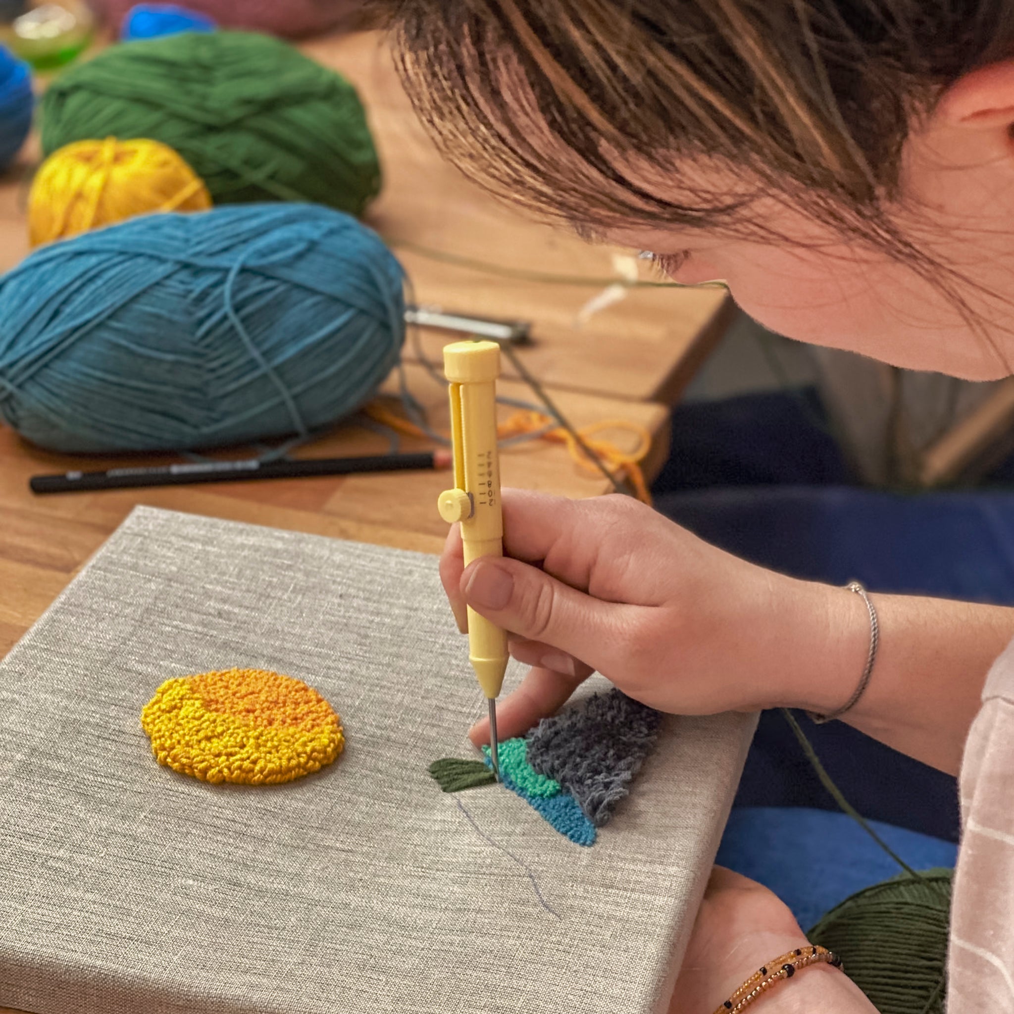 Fokus auf die Führung eines gelben Punch-Needle-Werkzeugs beim Gestalten einer sonnigen Berglandschaft auf gespanntem Textil.