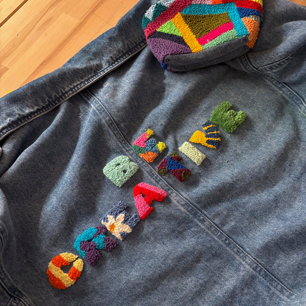 Jeansjacke auf Holzhintergrund mit bunten Punch-Needle-Buchstaben und geometrischen Patches.