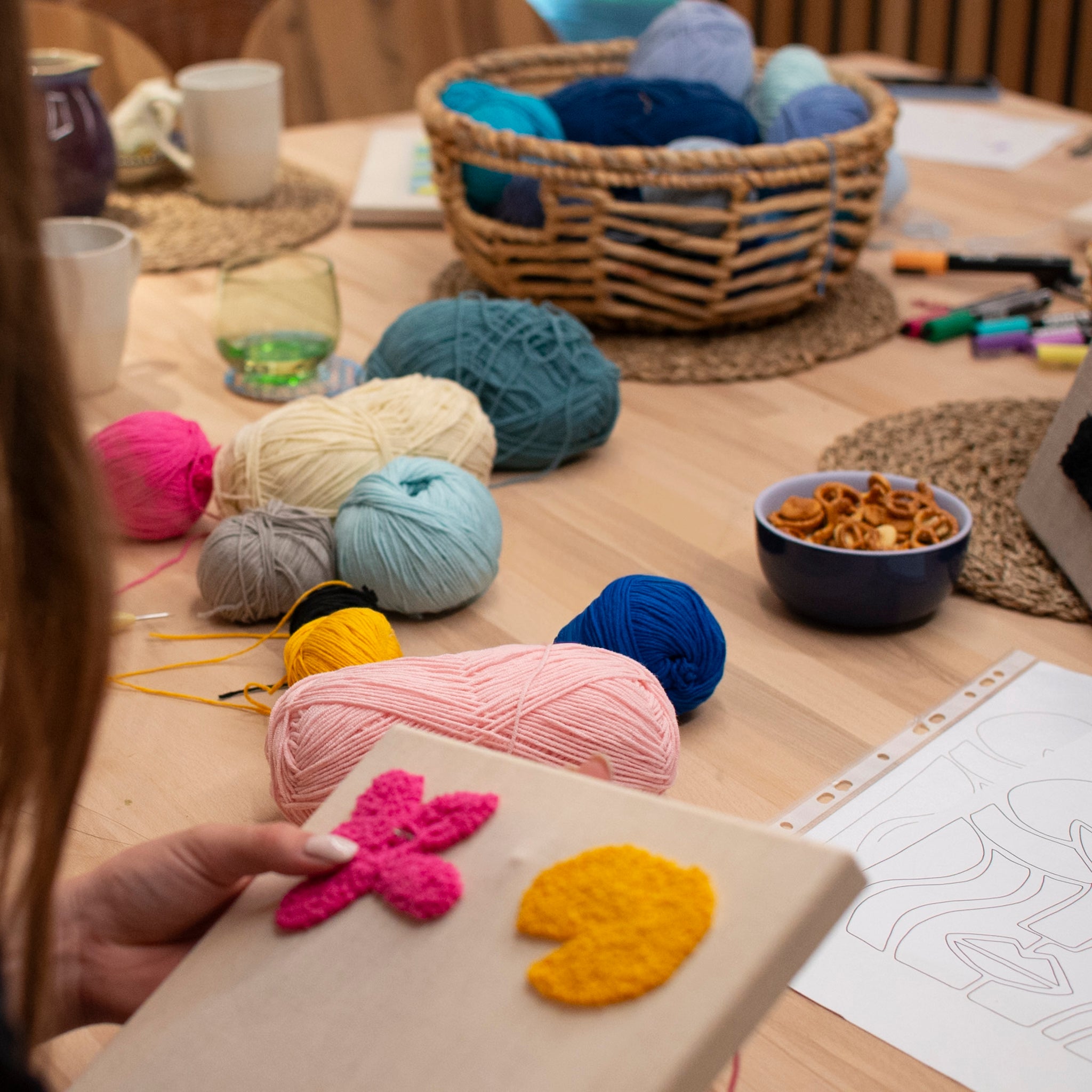 Blick auf einen Workshoptisch mit bunten Wollknäueln, Vorlagen, Snacks und angefangenen Projekten in gemütlicher Atmosphäre.