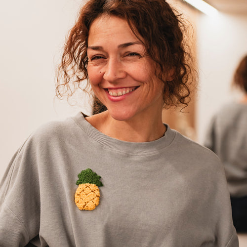 Porträt von Hülya Pfisterer mit einem grauen Sweatshirt, das durch einen gelben Ananas-Punch-Needle-Patch veredelt wurde.