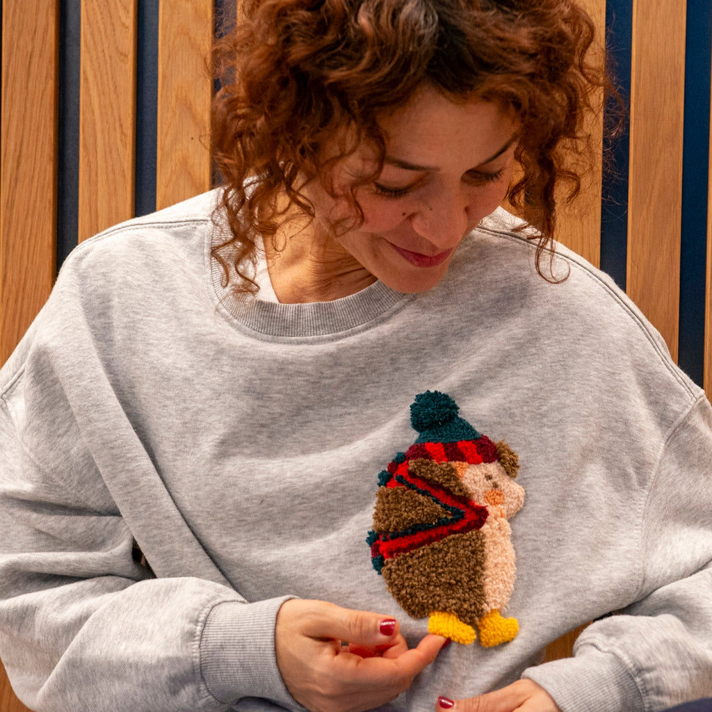 Lächelnde Hülya Pfisterer trägt ein graues Sweatshirt mit einem handgefertigten Igel-Patch in Punch-Needle-Technik.