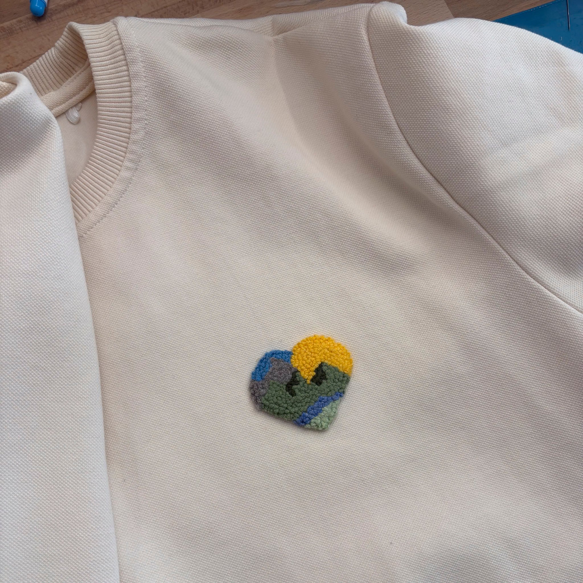 Nahaufnahme eines weißen Sweatshirts mit einem kleinen, herzförmigen Landschafts-Patch in Punch-Needle-Optik.