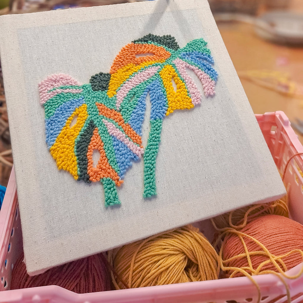 Abstraktes Monstera-Blatt in bunter Punch-Needle-Technik auf Leinwand, dekorativ präsentiert in einem Korb mit Wollknäueln.