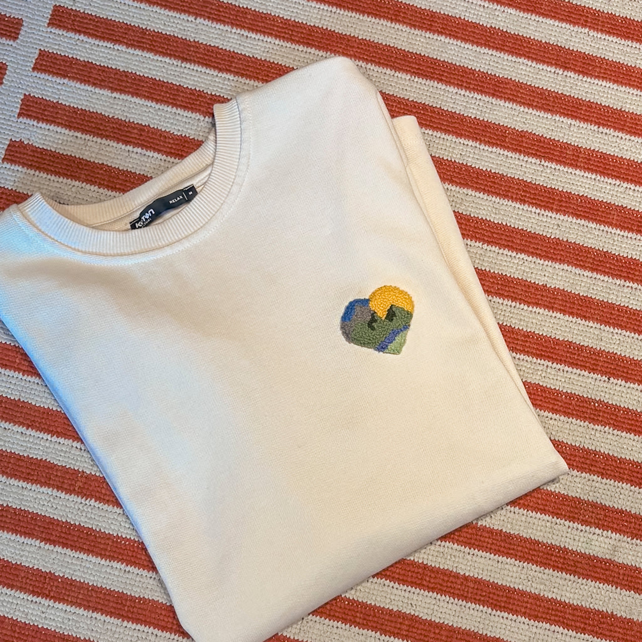 Flach liegendes weißes Sweatshirt mit einem dezenten Herz-Punch-Needle-Patch auf einem gestreiften Teppich-Hintergrund.