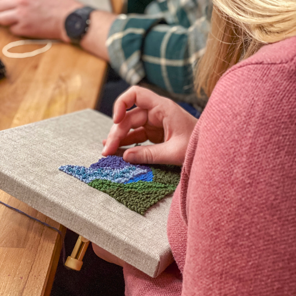 Nahaufnahme einer Teilnehmerin, die haptische blaue und lila Schlaufenstrukturen einer Berglandschaft auf eine Leinwand stickt.