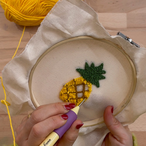 Hände füllen ein Ananas-Motiv im Stickrahmen mit einer Punch Needle, Fokus auf die handwerkliche Technik des Punch Needling