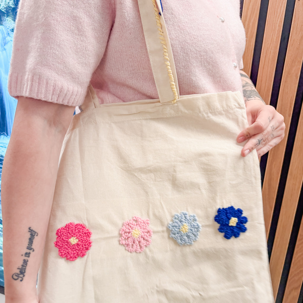 Eine Person in einem rosa Oberteil hält eine StraightCurls-Tragetasche mit vier Punch-Needle-Blumen in der Hand, die im TIM-Workshop in Augsburg angefertigt wurden.