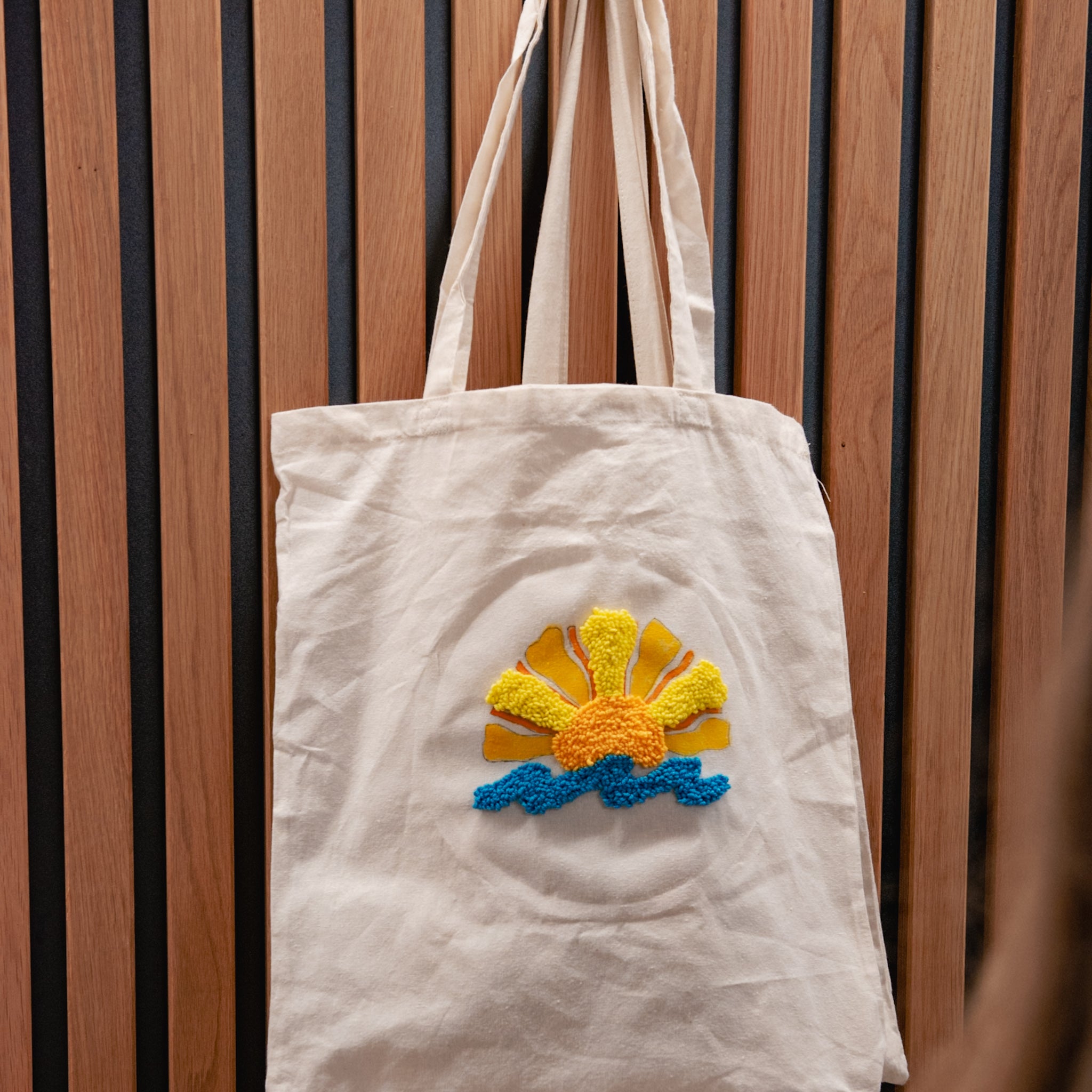 „StraightCurls Punch Needle“-Jutetasche von TIM Augsburg, weiße Tragetasche mit Motiv aus gelber Sonne und blauen Wellen.