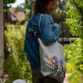 Person im Freien in einer blauen Jeansjacke mit Aufnähern, die einen „StraightCurls Punch Needle Jutebeutel“ (TIM Augsburg) in der Hand hält.