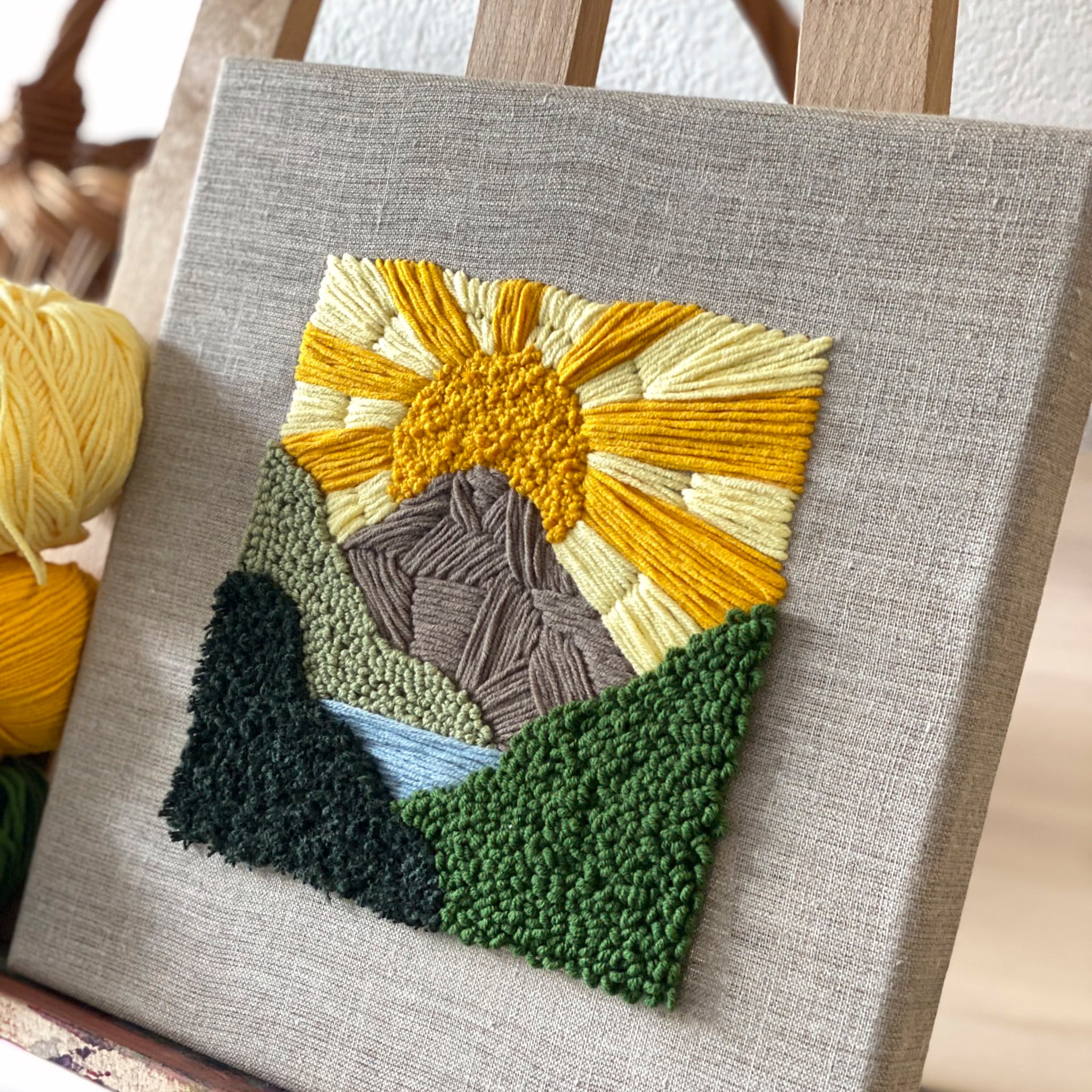 Quadratisches textiles Kunstwerk einer strahlenden gelben Sonne über grünen Bergen, gefertigt in strukturierter Punch-Needle-Manier.