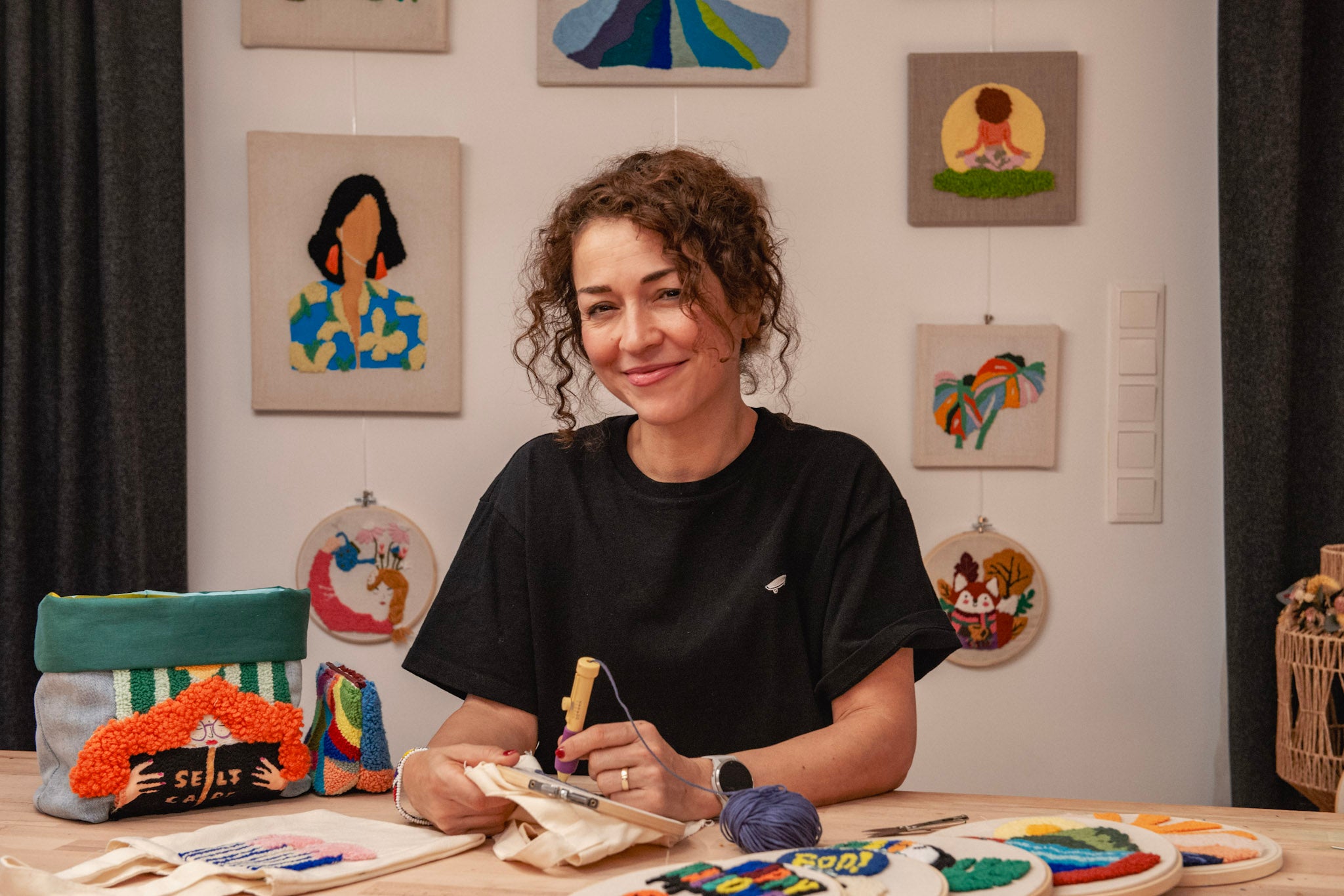 Hülya Pfisterer von StraightCurls beim Punch Needling im Atelier in Emmering bei Fürstenfeldbruck, DIY-Sets auf dem Tisch und fertige Punch Needle Motive im Hintergrund