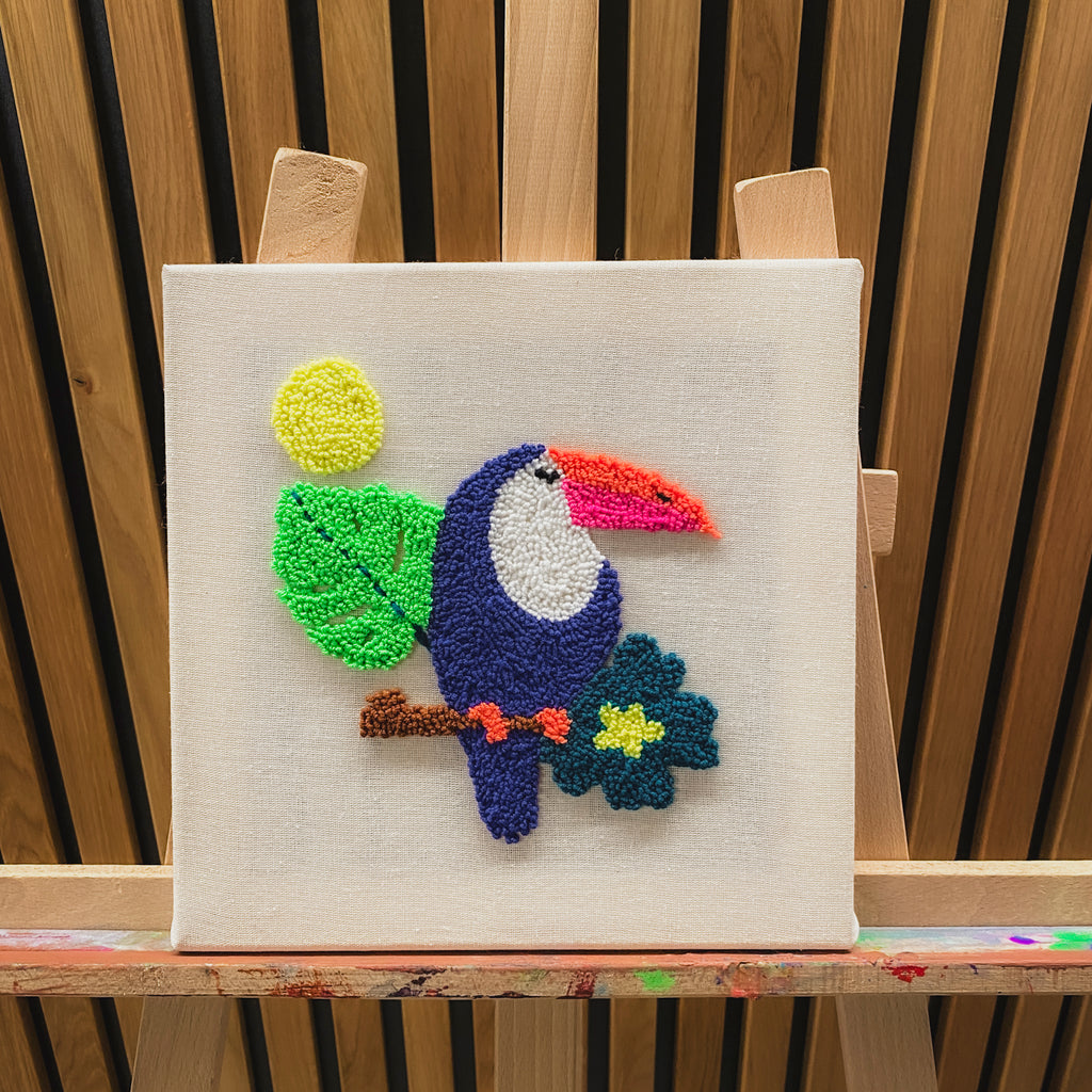 Farbgewaltiges Wandbild eines Tukans auf einem Ast, handgefertigt in plastischer Punch-Needle-Struktur auf einer Leinwand.