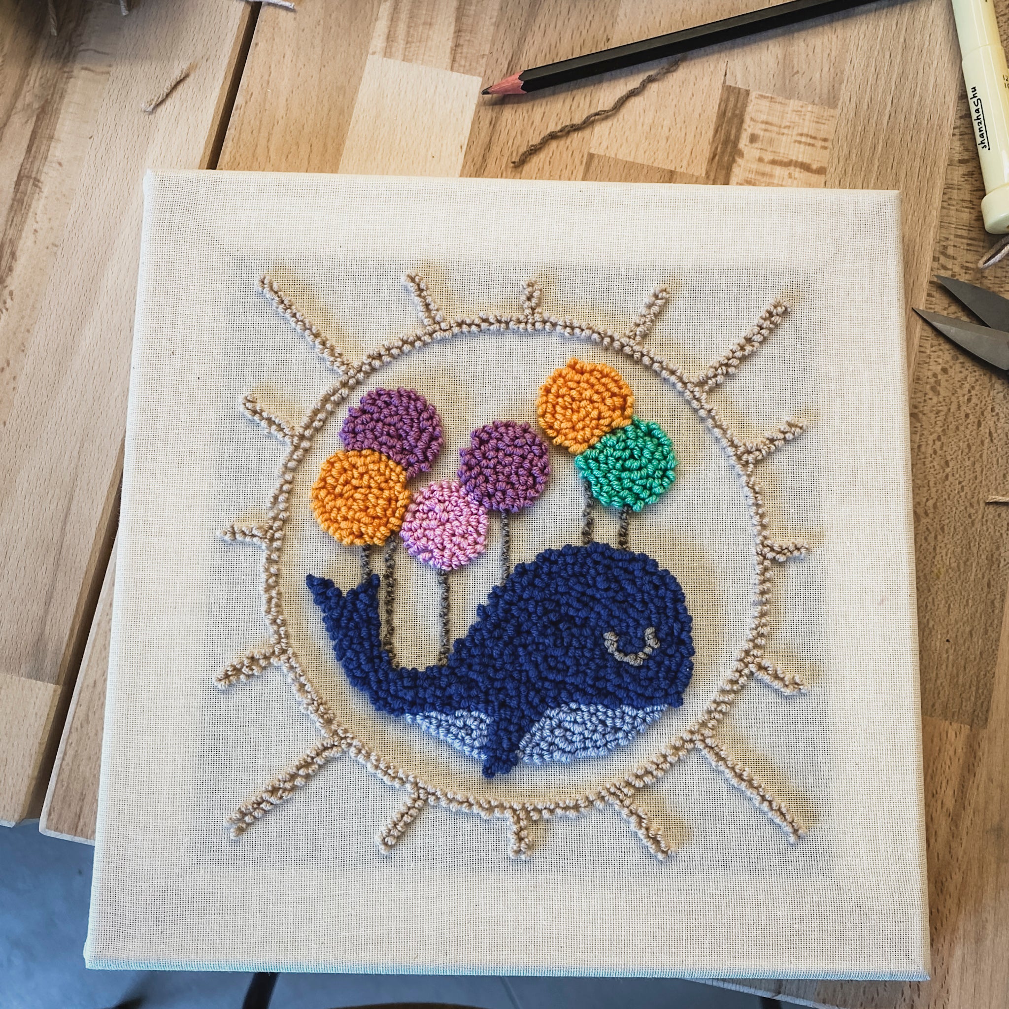 Handgefertigtes Wandbild eines blauen Wals mit bunten Luftballons, umgeben von einem gestickten Strahlenkranz in haptischer Optik.