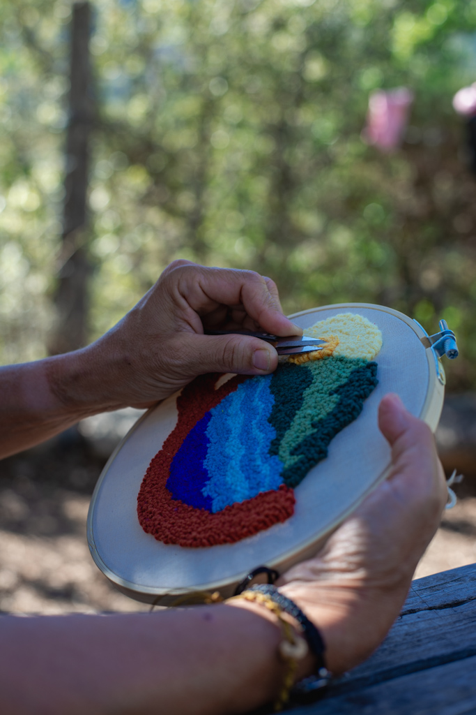 Frau hält Stickrahmen mit Punch Needle Motiv, rote Tasse mit blauen Wellen, Bergen und gelber Sonne, Handarbeit im Freien – kreative workshops muenchen FFB