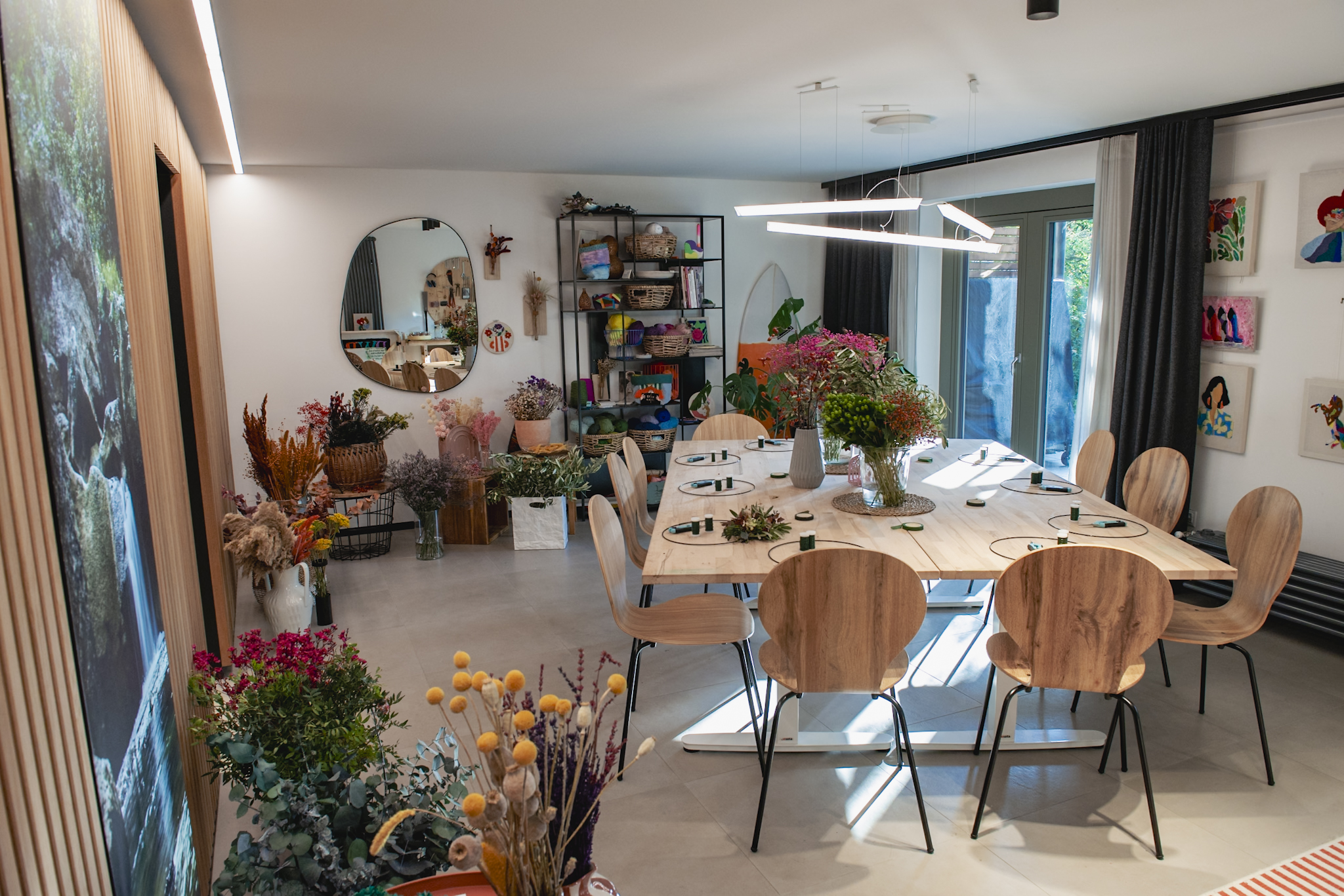 Impressionen aus dem Blumenkranz-Workshop im StraightCurls Atelier in Fürstenfeldbruck Emmering: Teilnehmerinnen binden an hellen Holztischen herbstliche Kränze aus frischen Blumen und Trockenblumen um einen Metallring. Man sieht Scheren, Draht und eine große Auswahl an Naturmaterialien in einer gemütlichen Workshop-Atmosphäre.